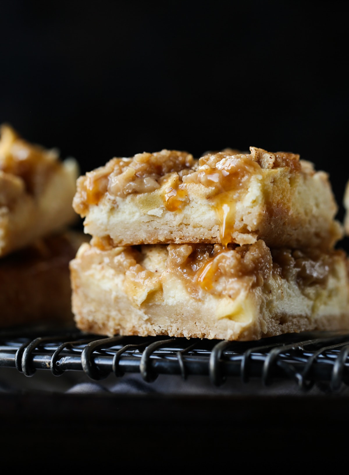 Caramel Apple Cheesecake Bars are creamy, sweet and delicious. They're a simple, buttery crust with an easy apple cheesecake filling topped with an oat streusel and caramel. 2 Apple Cheesecake Bars stacked on a wire rack