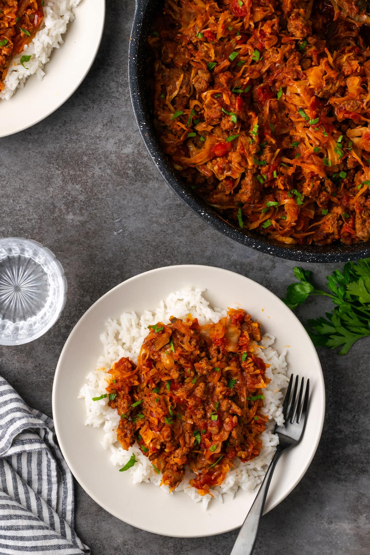 This easy unstuffed cabbage recipe is a savory one-pot meal made with juicy ground beef, pork, tangy fire-roasted tomatoes, and onions. Overhead view of unstuffed cabbage served over rice on a white plate next to more cabbage in a large pot.