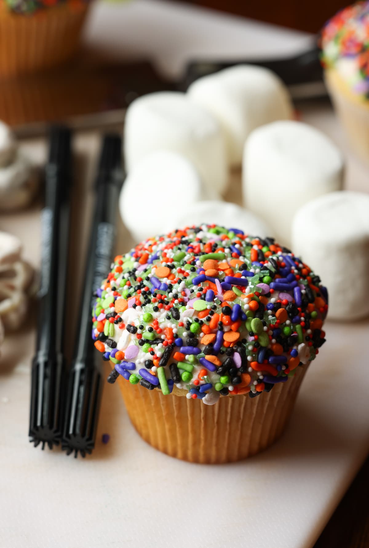 These are adorable Skeleton Halloween Cupcakes that are perfect for spooky season! Easy to make and so cute for classroom parties and a fun activity to do with your kids! Cupcake covered in Halloween themed sprinkles on a cutting board surrounded by marshmallows and edible food markers