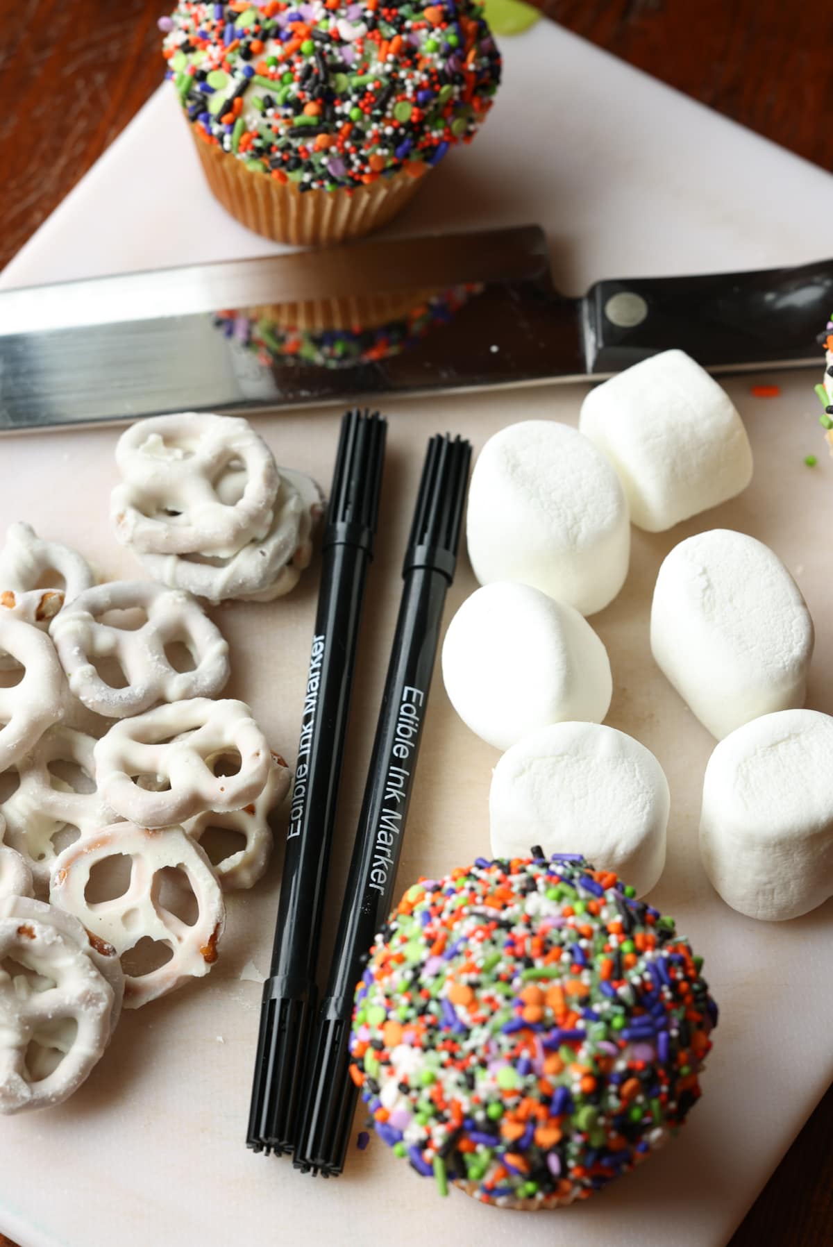 These are adorable Skeleton Halloween Cupcakes that are perfect for spooky season! Easy to make and so cute for classroom parties and a fun activity to do with your kids! Marshmallows, white chocolate covered pretzels, food coloring markers, and cupcakes on a white cutting board