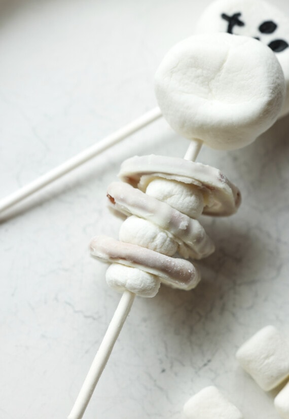 A lollipop stick threaded with mini marshmallows and white chocolate covered pretzels making edible skeleton decorations