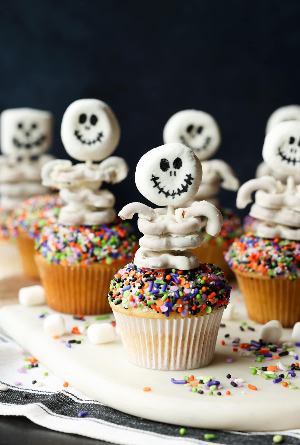 These are adorable Skeleton Halloween Cupcakes that are perfect for spooky season! Easy to make and so cute for classroom parties and a fun activity to do with your kids! Skeletons with marshmallow heads and white chocolate pretzel "rib cages" on top of sprinkle covered cupcakes