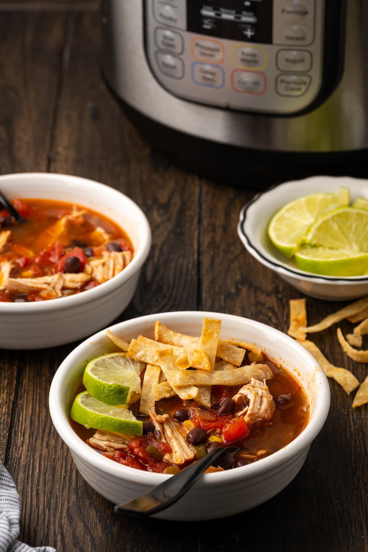 Smoky and flavorful Instant Pot chicken tortilla soup is a comforting one-pot meal that's easy to make any night of the week! A bowl of chicken tortilla soup garnished with lime wedges and crispy tortilla strips, with a bowl of lime wedges in the background next to the Instant Pot.