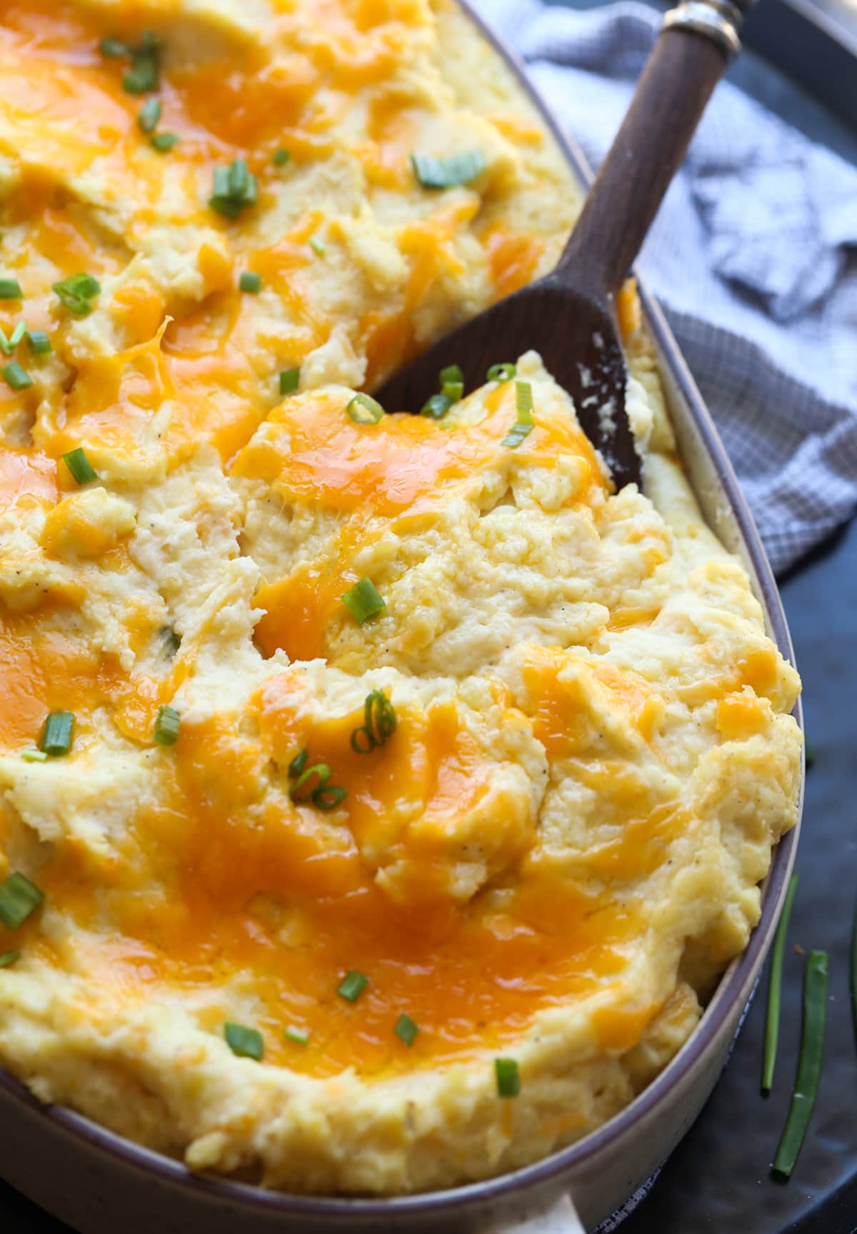These Cheesy Baked Mashed Potatoes is the BEST mashed potato recipe ever! Mashed Potatoes topped with cheddar cheese in a casserole dish with a serving spoon