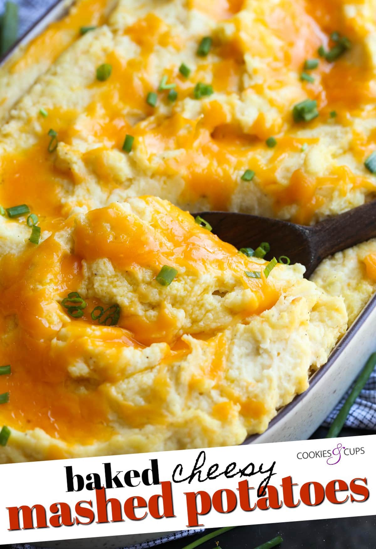 These Cheesy Baked Mashed Potatoes is the BEST mashed potato recipe ever! Scooping out a serving of cheesy mashed potatoes from a ceramic dish with writing on the image for Pinterest.