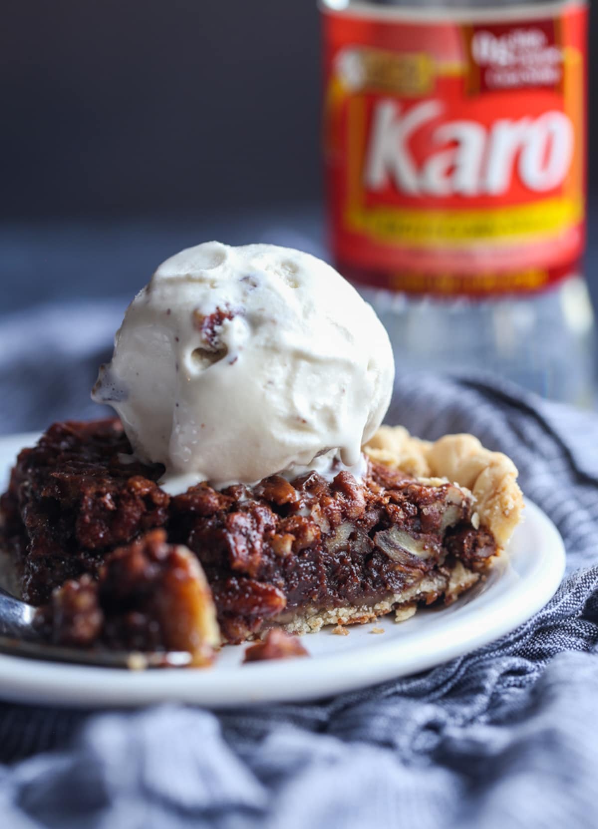 My EASY Chocolate Pecan Pie Recipe is a classic pecan pie recipe made even more delicious with melted chocolate, which turns the filling into a rich pudding-like texture loaded with pecans! A slice of chocolate pecan pie on a white plate with ice cream and a bottle of Karo syrup in the background
