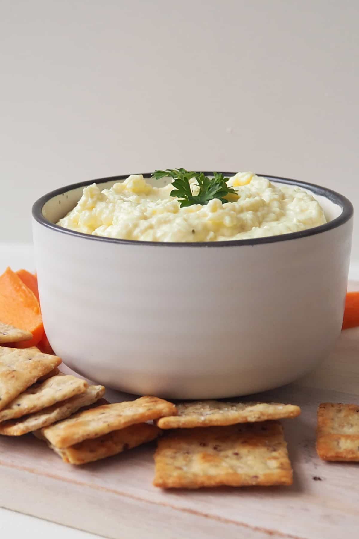Front view of French onion dip in a white bowl with dark striped rim that is sitting on a wooden serving platter with carrot sticks and crackers.