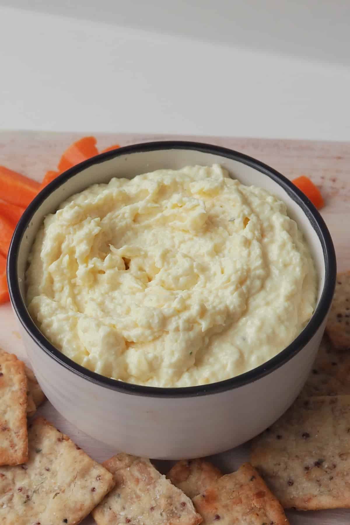 French onion dip in a white bowl with dark striped rim that is sitting on a wooden serving platter with carrot sticks and crackers.
