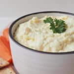 Side view of French onion dip in a white bowl with dark striped rim that is sitting on a wooden serving platter with carrot sticks and crackers.