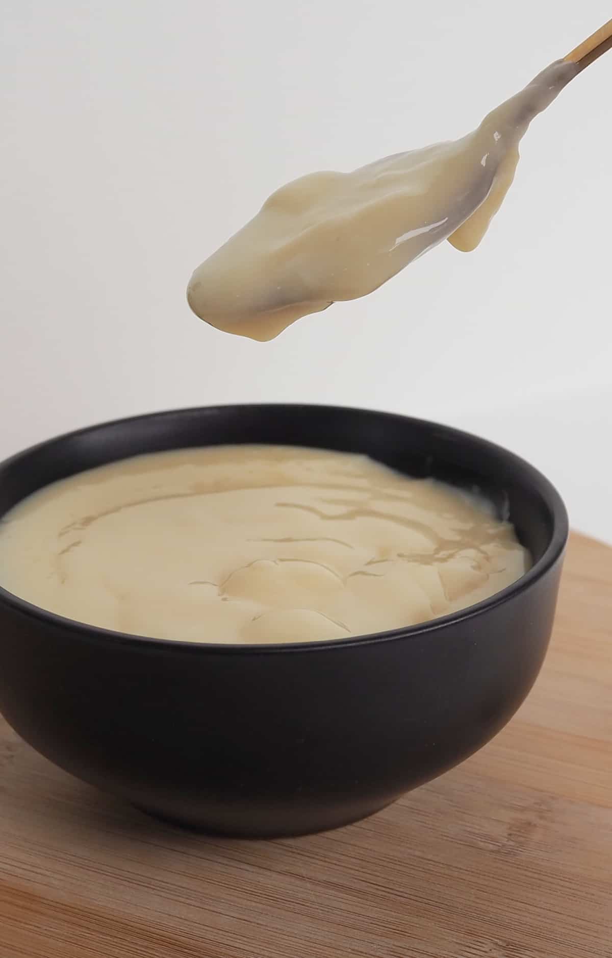 Brandy Custard in a black bowl sitting on a wooden board. A gold spoon with custard on it is above the bowl.