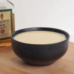Side view of Brandy Custard in a black bowl sitting on a wooden board. In the background is a bottle of brandy.