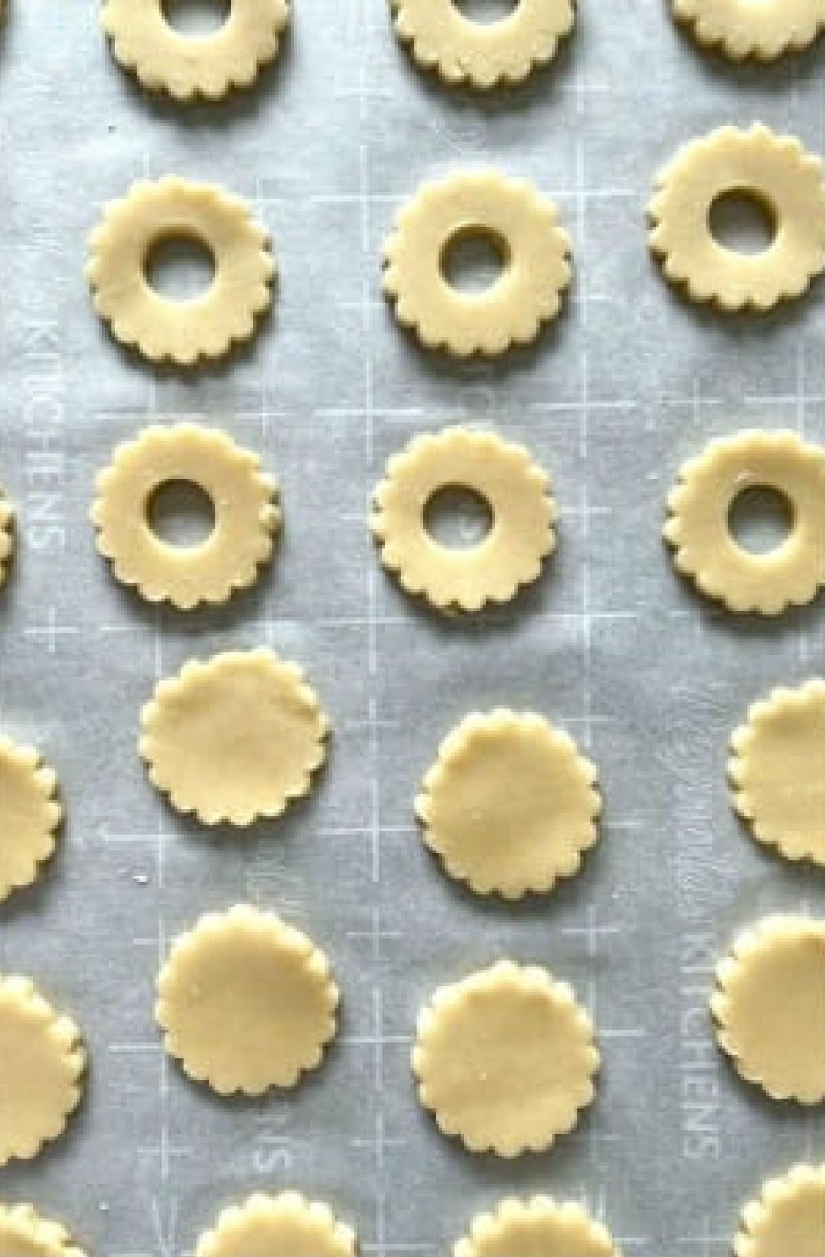 This is the absolute BEST Linzer Cookies recipe ever! These gorgeous and easy cookies are buttery, flaky shortbread filled with sweet jam and dusted with powdered sugar. A Christmas Cookie Classic! Cookie Dough cit out into scalloped circles and scalloped circles with a hole on a parchment lined baking sheet