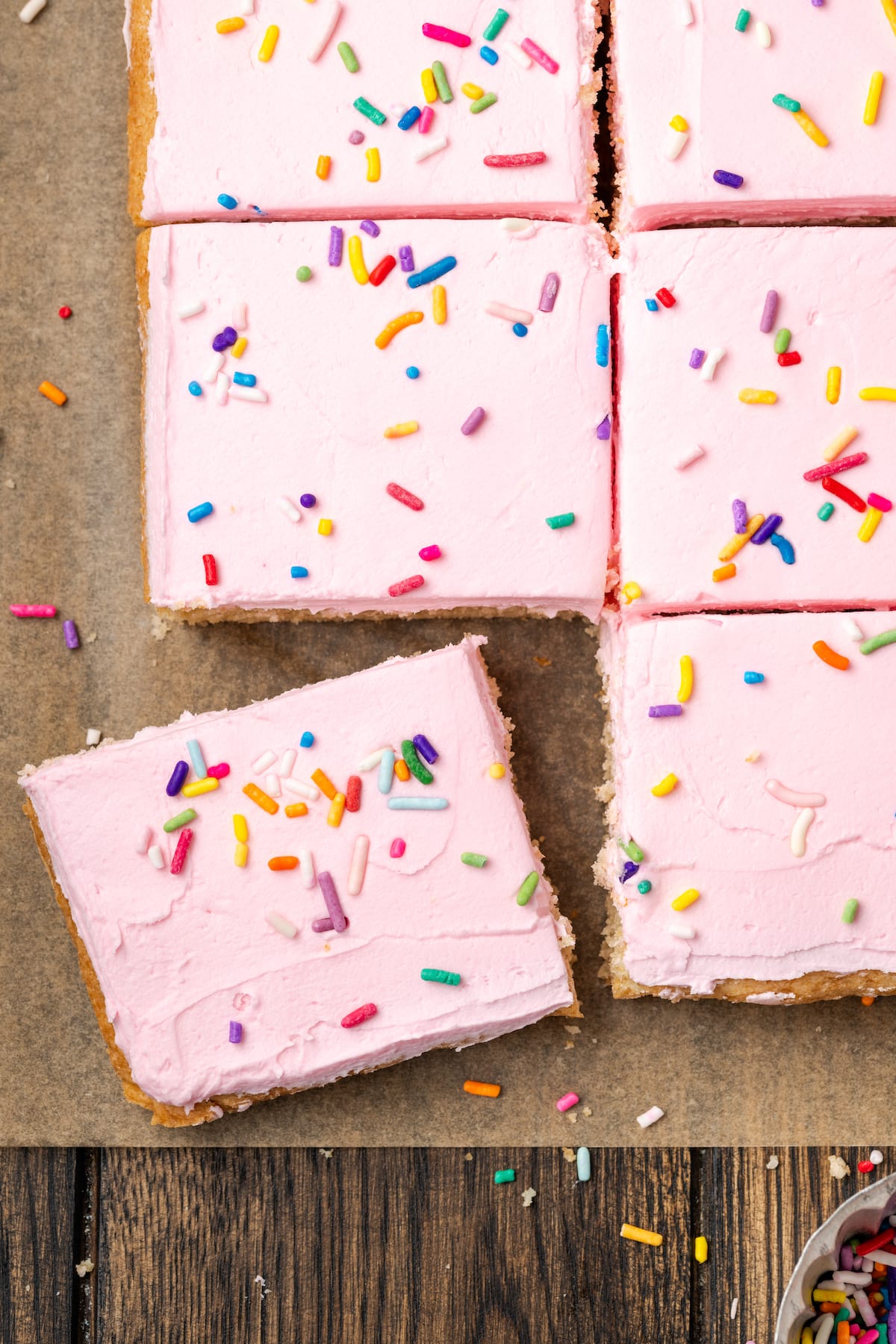 These soft and chewy frosted sugar cookie bars are topped with a thick layer of buttercream frosting (and sprinkles!). Ready in 30 minutes! Overhead view of frosted sugar cookie bars topped with rainbow sprinkles cut into squares on a parchment-lined cutting board.