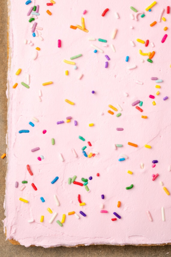 These soft and chewy frosted sugar cookie bars are topped with a thick layer of buttercream frosting (and sprinkles!). Ready in 30 minutes! Sugar cookie bars frosted with pink buttercream frosting and topped with rainbow sprinkles.