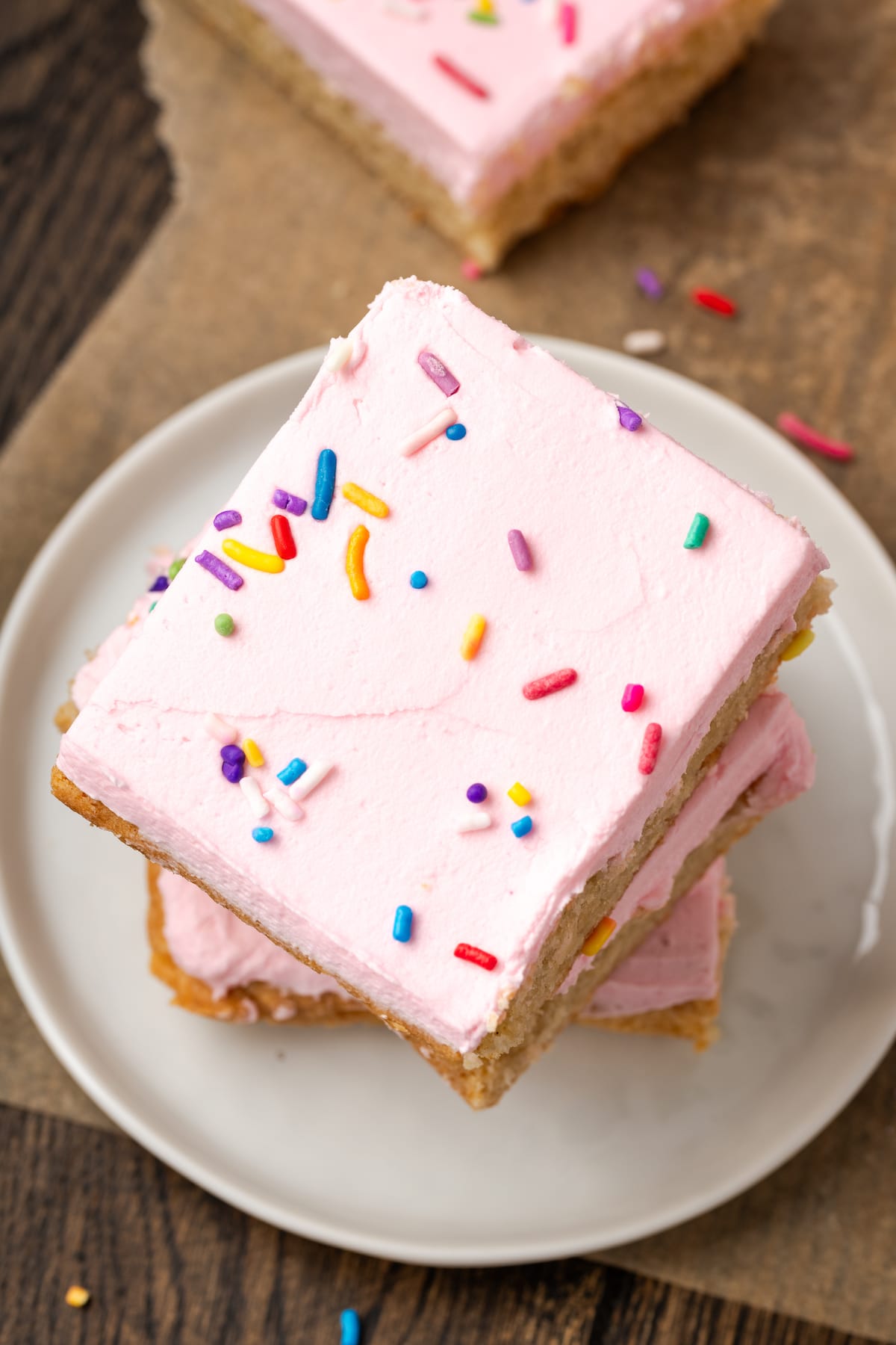 These soft and chewy frosted sugar cookie bars are topped with a thick layer of buttercream frosting (and sprinkles!). Ready in 30 minutes! Overhead view of three frosted sugar cookie bars stacked on a white plate.