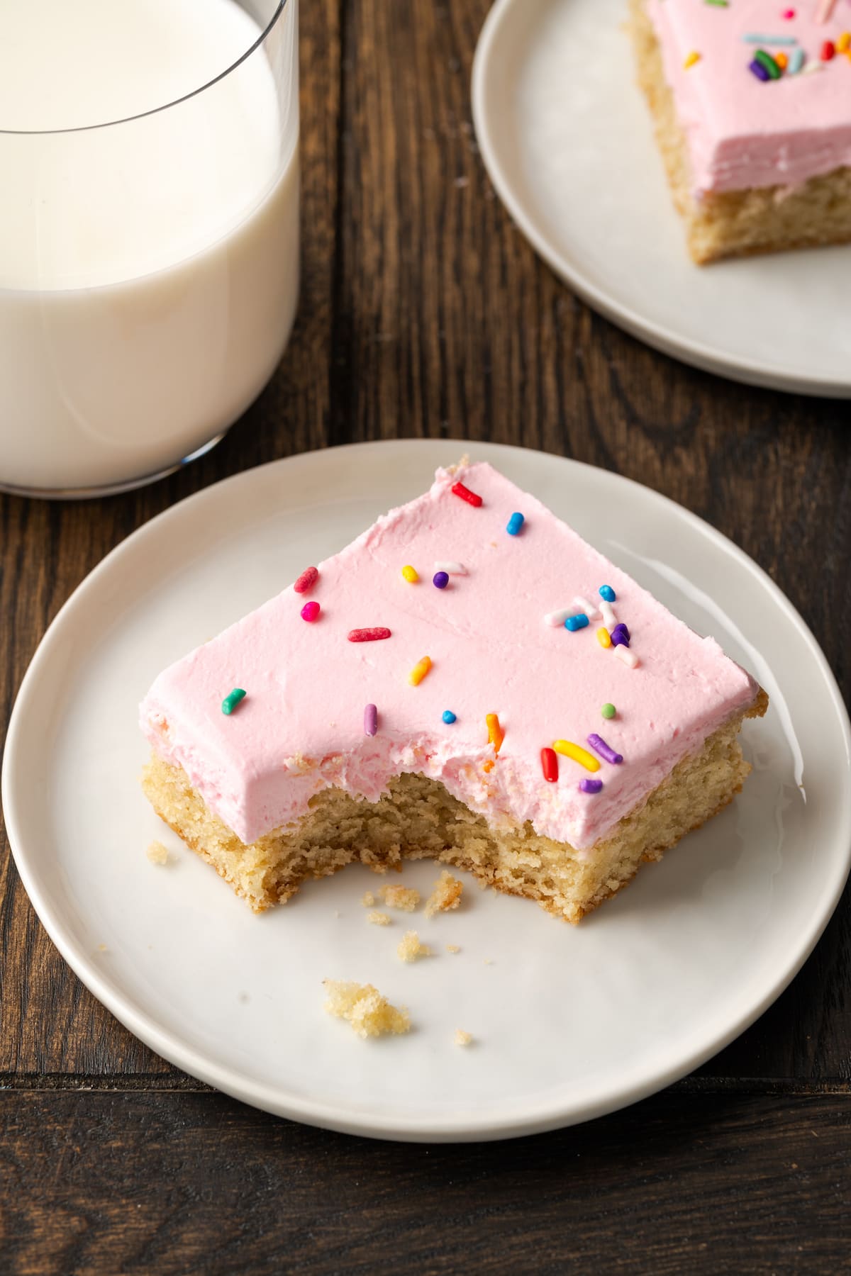 These soft and chewy frosted sugar cookie bars are topped with a thick layer of buttercream frosting (and sprinkles!). Ready in 30 minutes! A frosted sugar cookie bar with a bite missing on a white plate, next to a glass of milk.