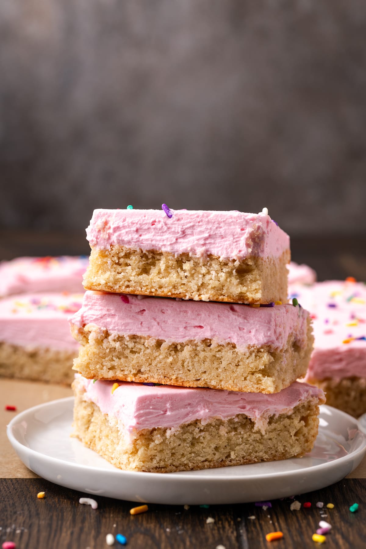 These soft and chewy frosted sugar cookie bars are topped with a thick layer of buttercream frosting (and sprinkles!). Ready in 30 minutes! Three frosted sugar cookie bars stacked on a white plate, with more bars in the background.