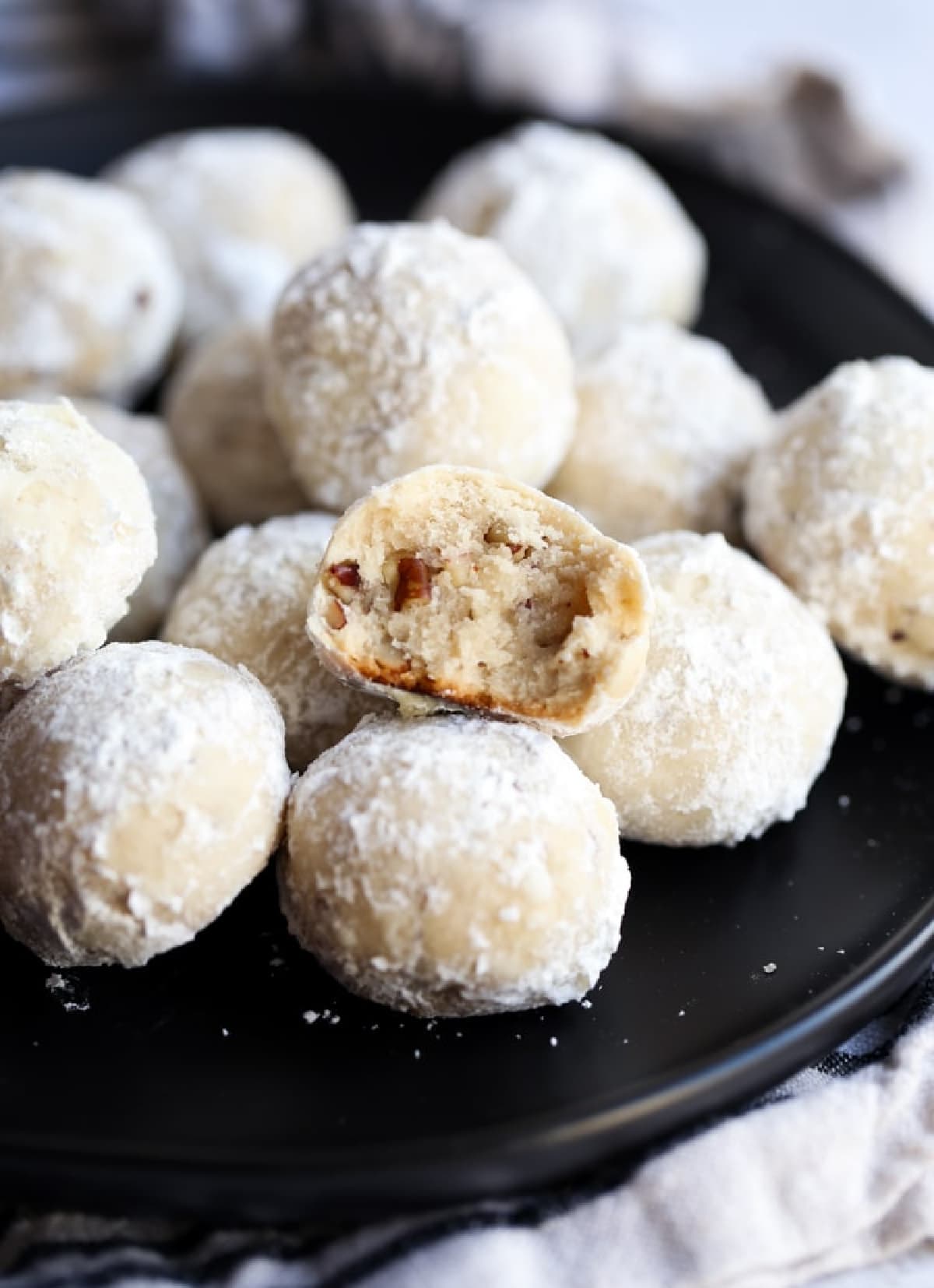 Russian tea cakes (also called snowball cookies) are rich and buttery holiday cookies coated in powdered sugar, filled with walnuts or pecans! A snowball cookie that is bitten in half on a black plate