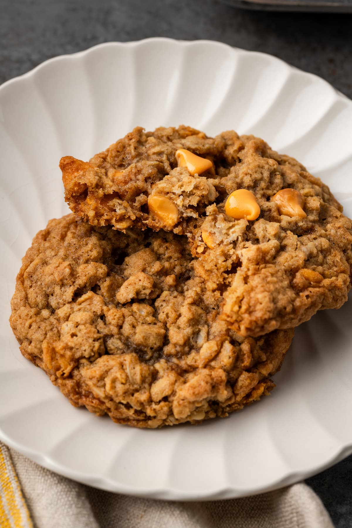 Soft and chewy oatmeal butterscotch cookies are rich and buttery, filled with tender oats, butterscotch morsels, and sweet cinnamon spice. Two oatmeal butterscotch cookies stacked on a white plate, with a bite missing from the top cookie.