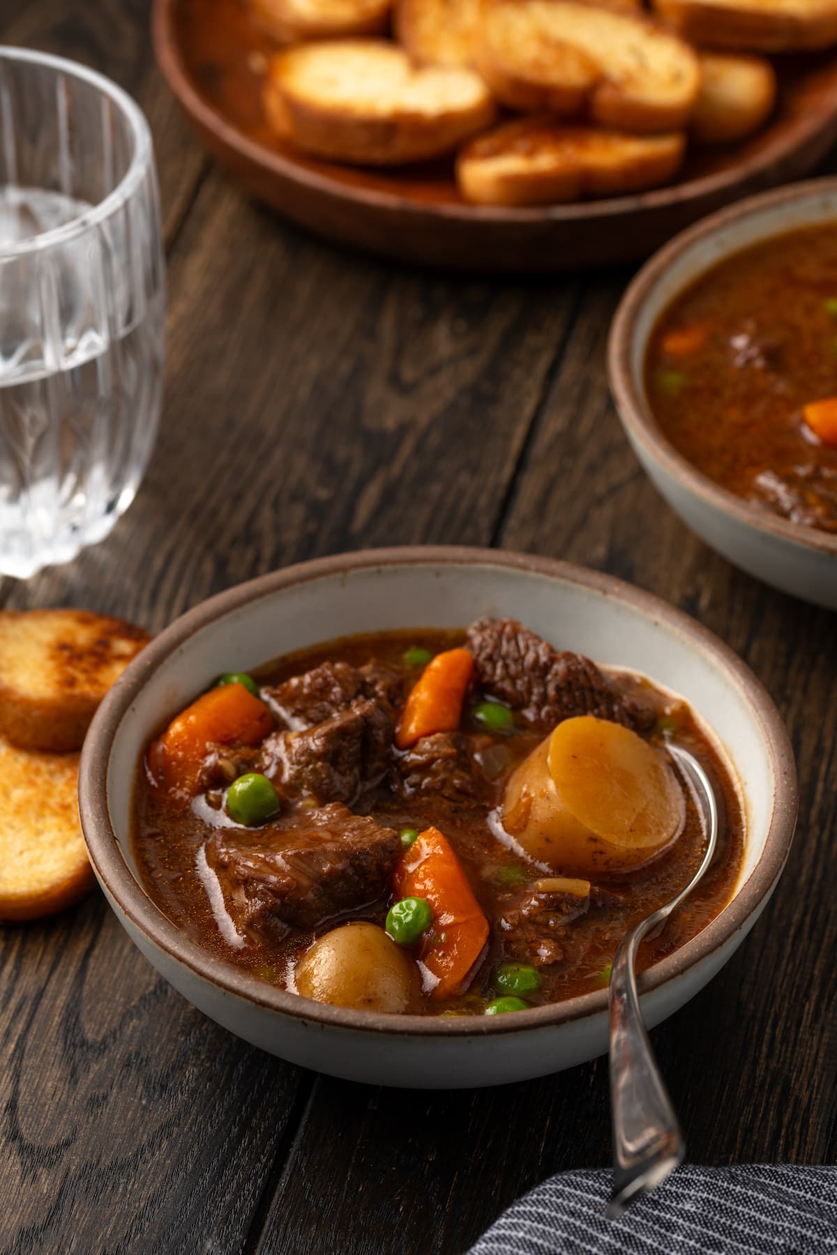 A bowl of Instant Pot beef stew on a wooden tabletop, with a plate of crostini in the background.