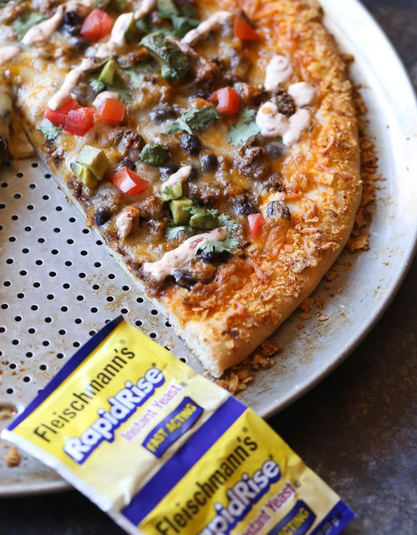 Taco pizza on a pizza pan with a slice missing, next to a yeast packet.
