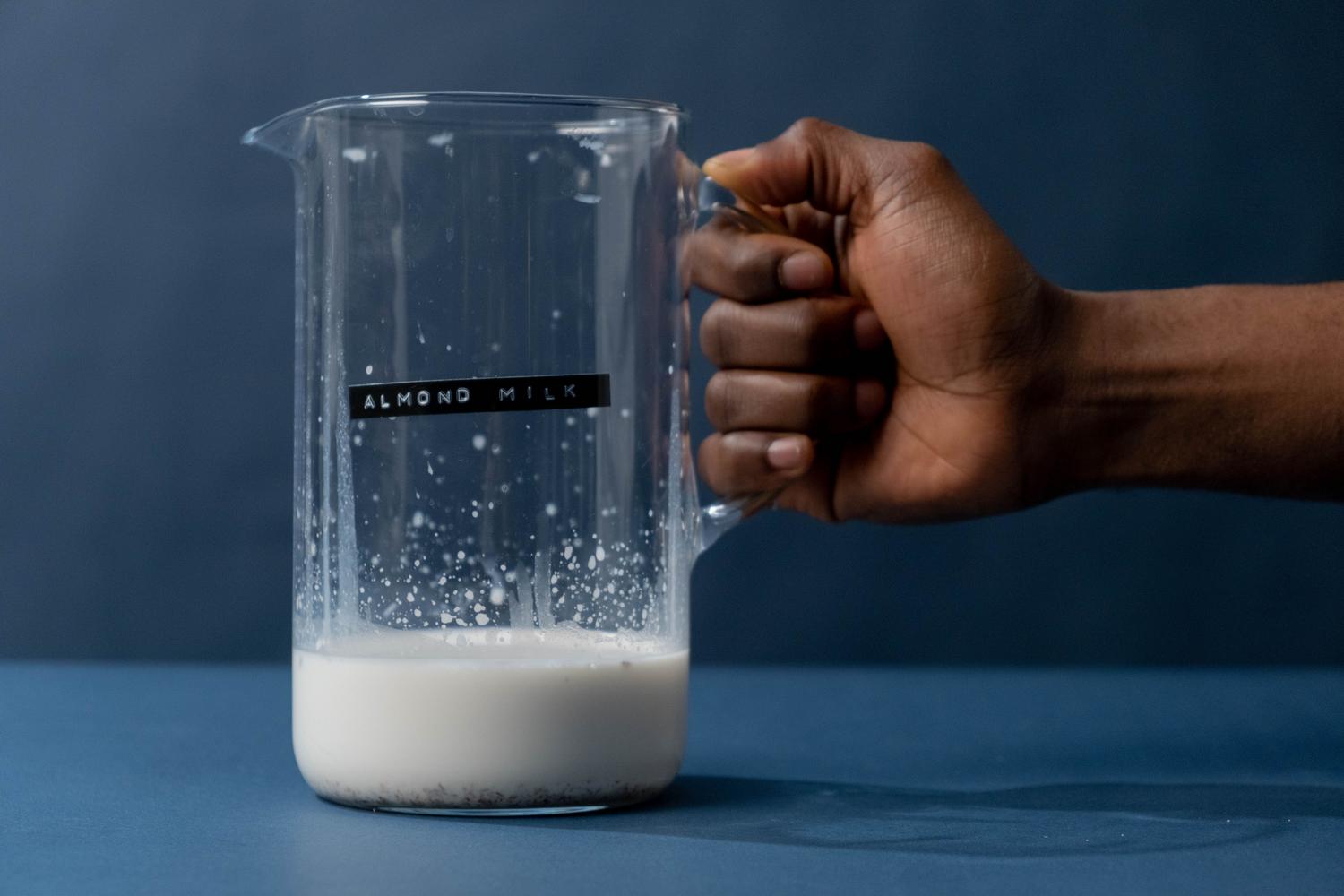 Primer plano de una mano vertiendo leche de almendras casera desde una jarra de cristal.