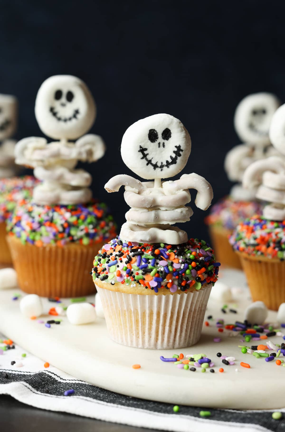 These are adorable Skeleton Halloween Cupcakes that are perfect for spooky season! Easy to make and so cute for classroom parties and a fun activity to do with your kids! Multiple sprinkle covered cupcakes decorated with skeleton toppers made from white chocolate covered pretzels.