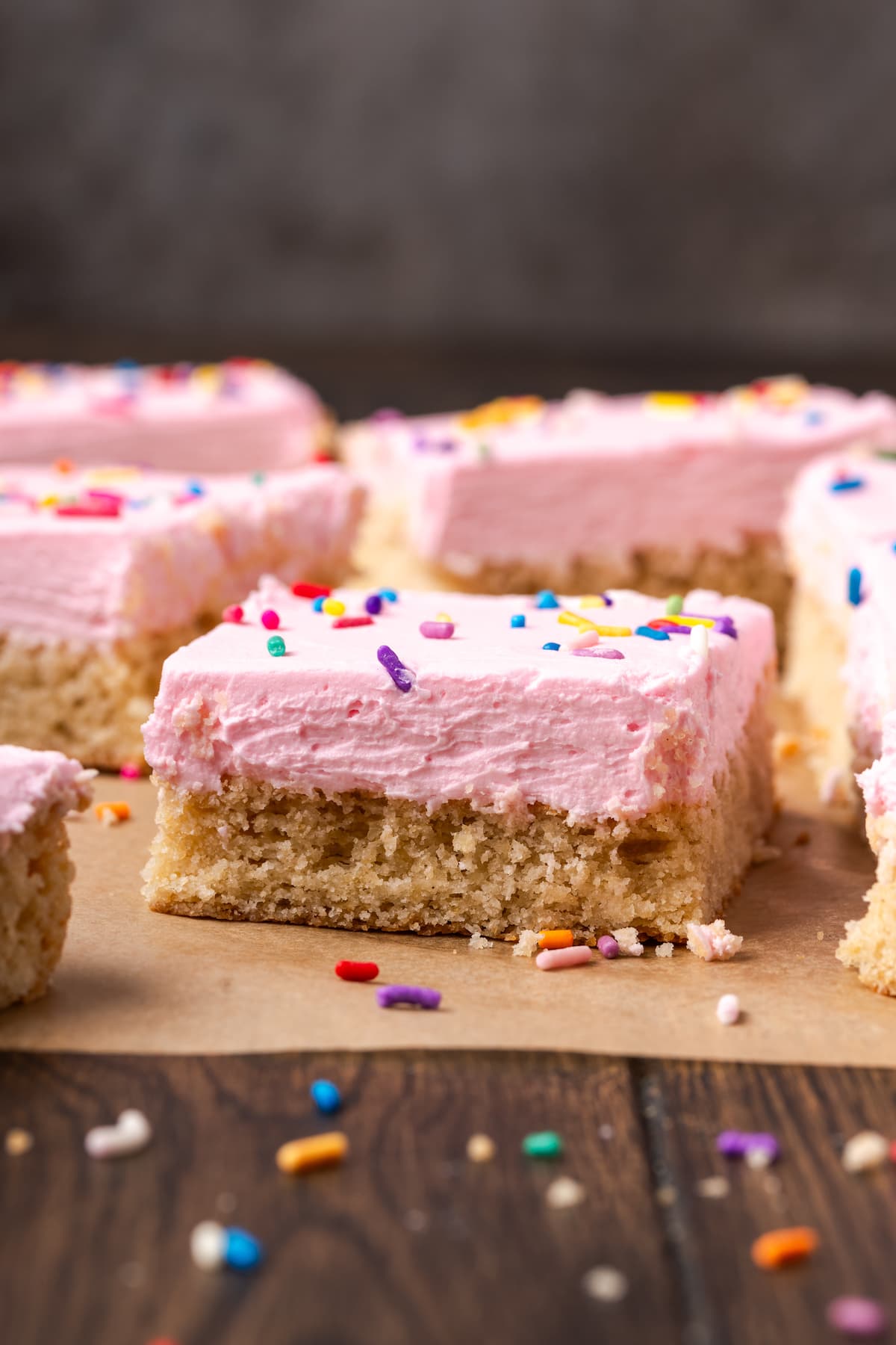 These soft and chewy frosted sugar cookie bars are topped with a thick layer of buttercream frosting (and sprinkles!). Ready in 30 minutes! Sugar cookie bars topped with pink frosting and rainbow sprinkles on a parchment-lined cutting board.