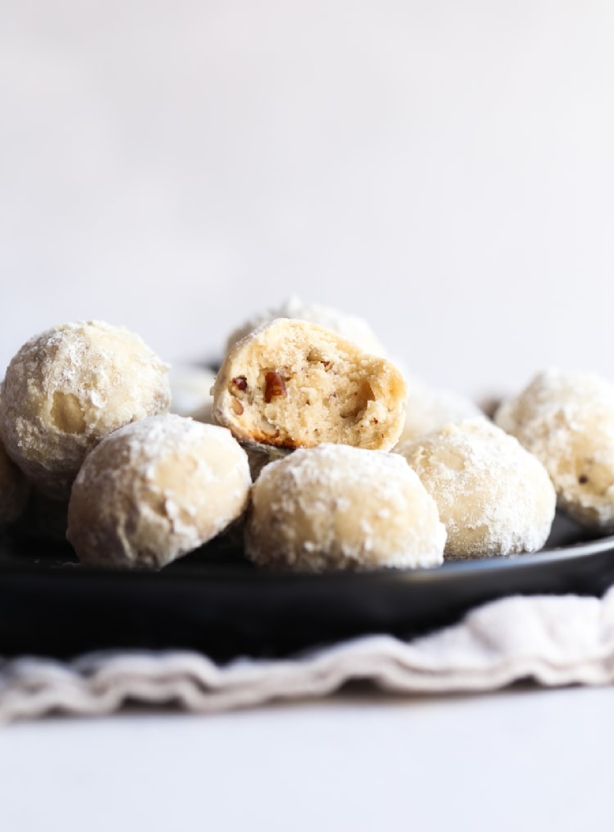 Russian tea cakes (also called snowball cookies) are rich and buttery holiday cookies coated in powdered sugar, filled with walnuts or pecans! Stacked Russian Tea Cakes on a black plate with one cut in half.