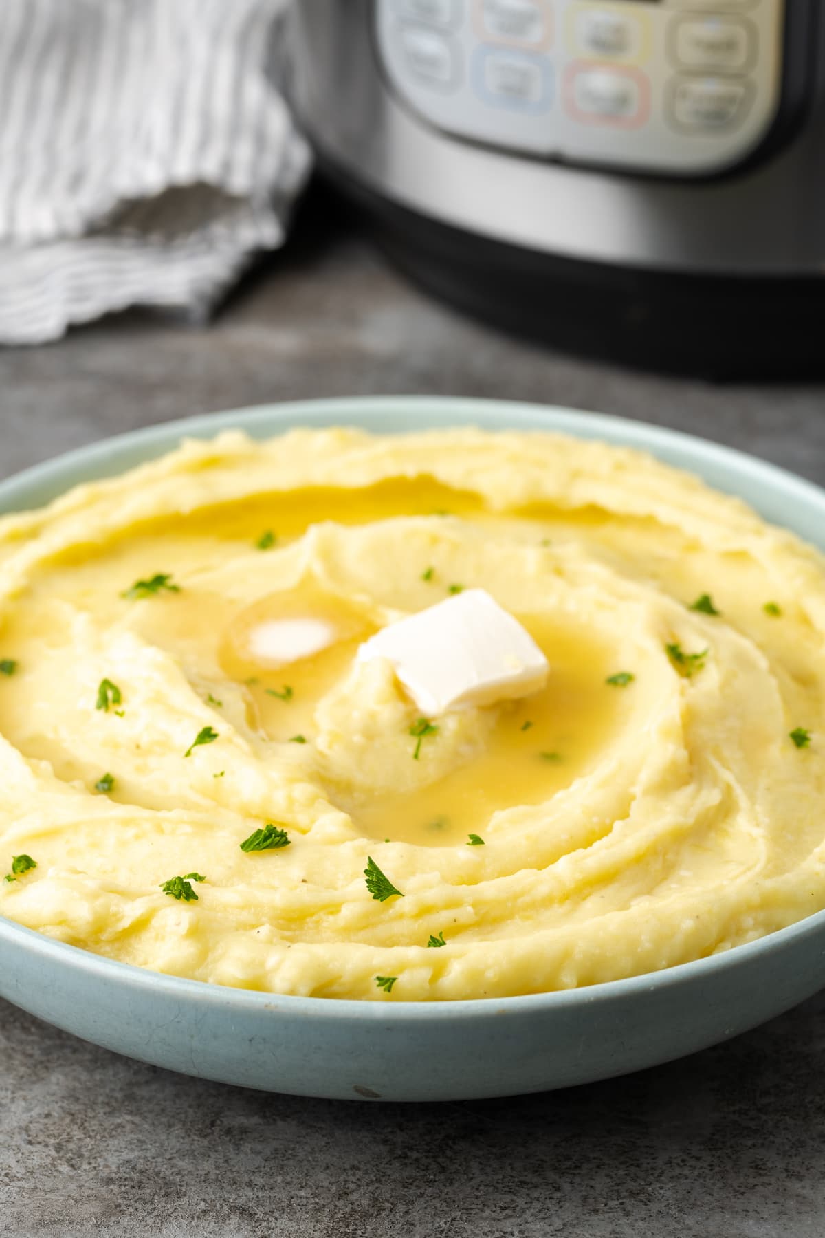 Buttery, fluffy Instant Pot Mashed Potatoes are a flavorful and easy side dish. Enjoy creamy homemade mashed potatoes, ready in half the time! Creamy Instant Pot mashed potatoes in a bowl topped with two pats of butter and chives, with the Instant Pot in the background.